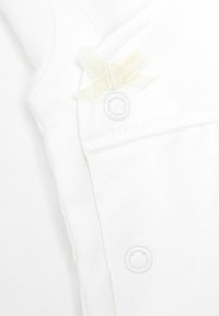 Vêtement en coton blanc avec un petit nœud décoratif crème et des boutons-pression. Texture douce avec une finition lisse et un design simple.