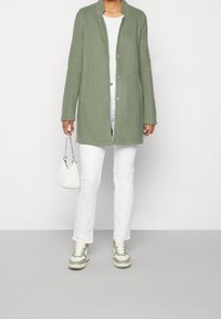 Persona che indossa un cappotto verde, una camicia bianca e pantaloni, tiene una borsa bianca, in piedi su un pavimento grigio con uno sfondo bianco.
