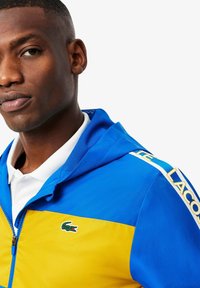 Blauwe en gele hoodie-jack met een rits, gaasstructuur, krokodillenlogo en "LACOSTE" in het geel bedrukt op de mouw.