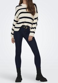 Gestrickter cremefarbener Pullover mit schwarzen Streifen, weiten Ärmeln, kombiniert mit dunklen Skinny-Jeans und schwarzen Stiefeletten. Ein goldener Gürtel betont die Taille.