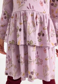 Dziecko w lawendowej sukience z kwiatowymi wzorami, z wieloma warstwami i długimi rękawami, z rękami swobodnie opartymi po bokach.