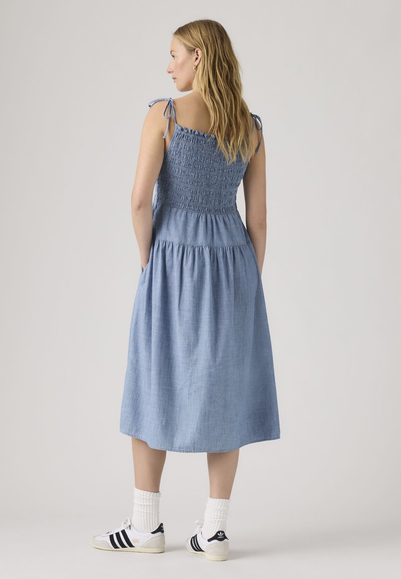 Femme aux longs cheveux portant une robe midi bleu sans manches avec des bretelles nouées, des chaussettes blanches et des baskets blanches à rayures noires, debout de dos.