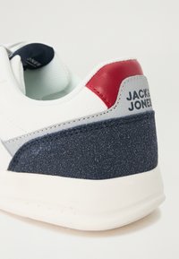 White sneaker featuring a red leather accent, a textured navy blue material at the heel, and brand logo on the side. Soft, rounded shape.