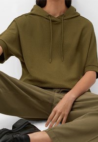 Sudadera de color verde oliva texturizada con mangas cortas y cordón, combinada con pantalones holgados a juego y zapatos negros de suela gruesa.