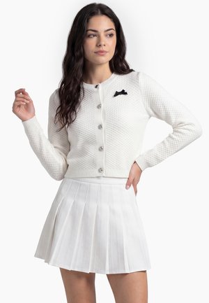 Donna che indossa un cardigan bianco con bottoni, dalla texture particolare, con un piccolo fiocco nero e una gonna bianca plissettata, posando con una mano sul fianco.