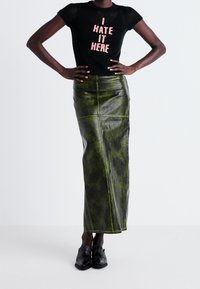 Camiseta negra de punto con el texto "ODIO ESTO AQUÍ" en color rosa, combinada con una falda larga de cuero verde con un acabado brillante y costuras visibles.