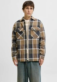 Camicia a quadri nei toni del marrone, beige e nero. Caratterizzata da due tasche sul petto, chiusura con bottoni e un tessuto morbido e texturizzato. Indossata sopra un maglione color crema.