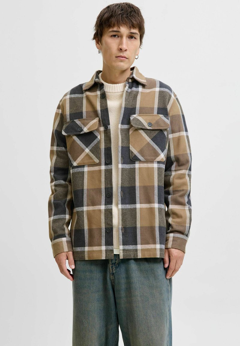 Camicia a quadri nei toni del marrone, beige e nero. Caratterizzata da due tasche sul petto, chiusura con bottoni e un tessuto morbido e texturizzato. Indossata sopra un maglione color crema.