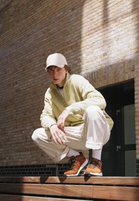 Chaqueta de cremallera verde claro, pantalones blancos y zapatillas marrones. Lleva una gorra beige, sentado en unos escalones de madera con un fondo de ladrillo.