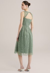 Blåsdklänning i dämpad grön färg med en åtsittande bodice, utsvängd kjol och intrikata blommönster. Har en korsad ryggdesign och en vågig fåll.
