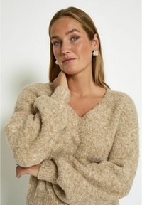 Beige gebreide trui met een V-hals en pofmouwen, voorzien van een zachte, gestructureerde afwerking. Zilveren ringoorbellen accentueren de look.