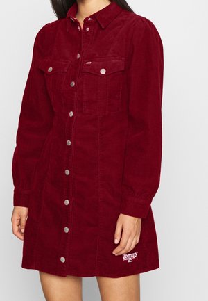 Femme portant une robe burgundy en velours côtelé à manches longues, boutonnée devant, avec poches poitrine et logo Tommy Jeans près de l’ourlet.