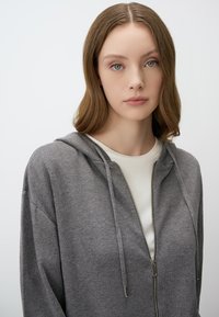 Haut à capuche zippé gris avec une texture douce, comportant une fermeture éclair à l'avant, un cordon de serrage à la capuche et une coupe décontractée, superposé sur un T-shirt blanc.