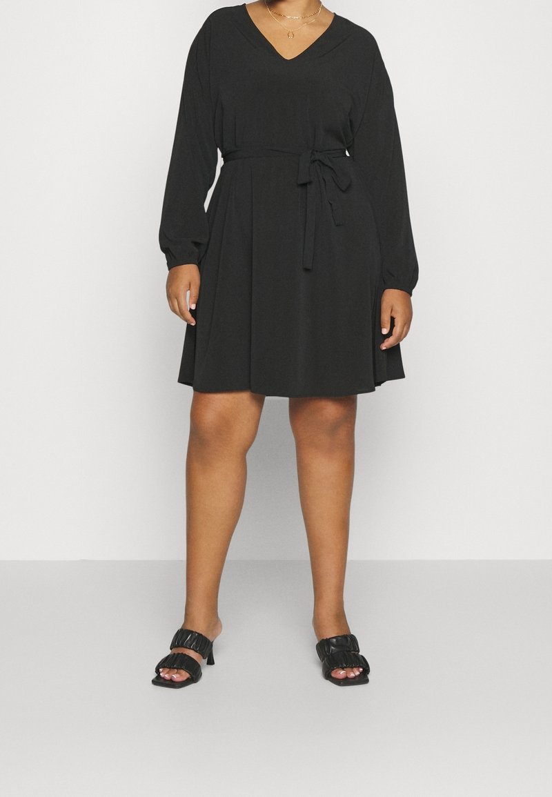 Femme portant une robe noire à manches longues avec une ceinture à la taille et des sandales noires à talons, debout devant un fond clair uni.