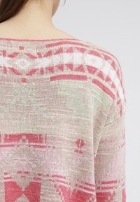 Lehký svetr s vzorem pleteniny, který má geometrický design v růžové a krémové barvě. Měkká textura, široký výstřih a vpředu padající ramena.