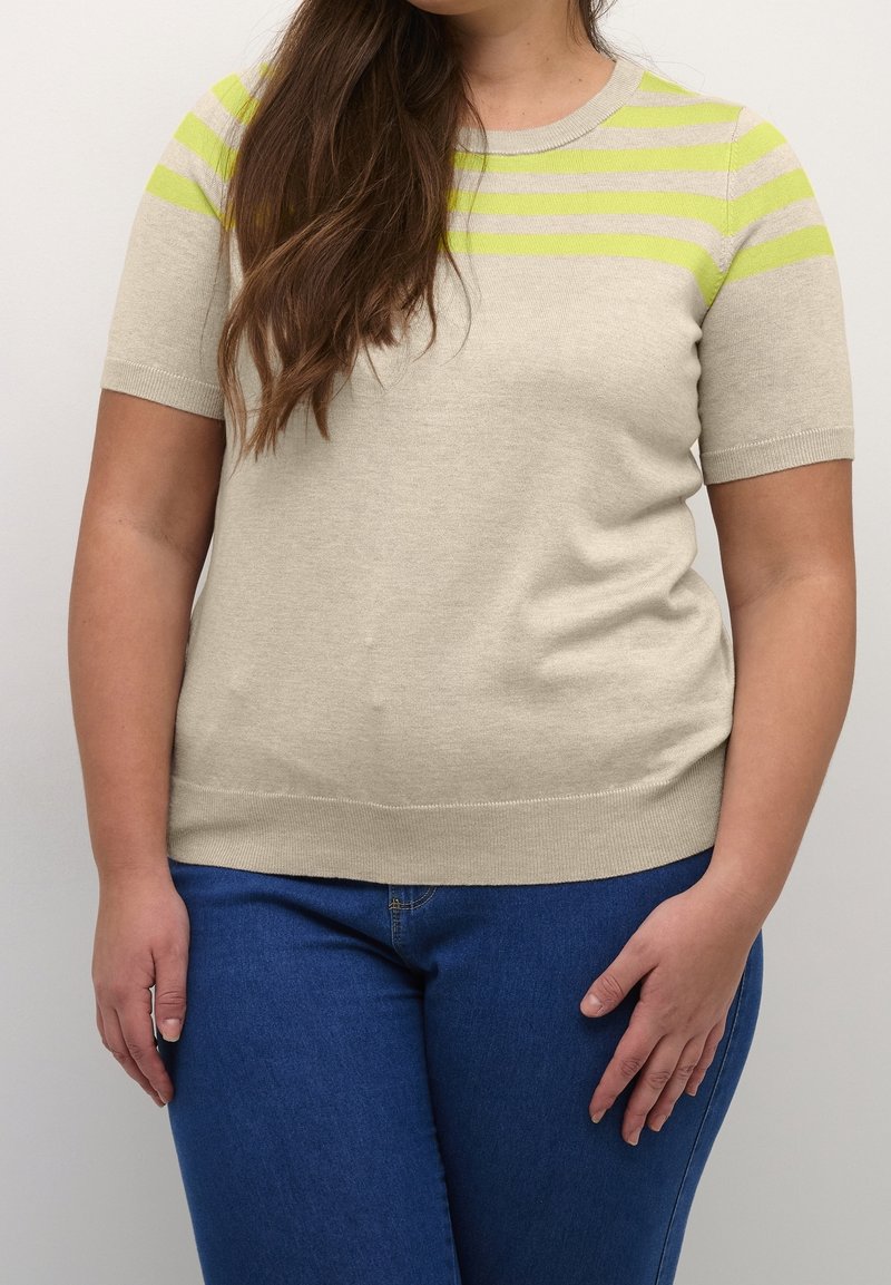Femme aux longs cheveux bruns porte un pull beige à manches courtes avec des rayures jaunes et un jean bleu sur un fond uni.