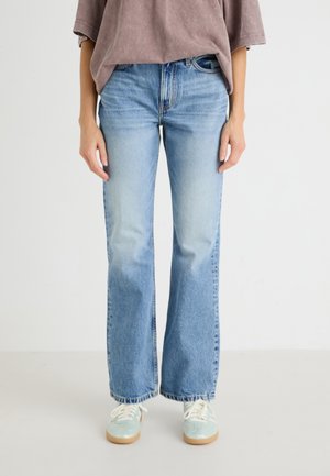 Person iført lyseblå jeans med lige ben, en løs brun top og lyseblå sneakers med hvide snørebånd, stående mod en ensfarvet hvid baggrund.