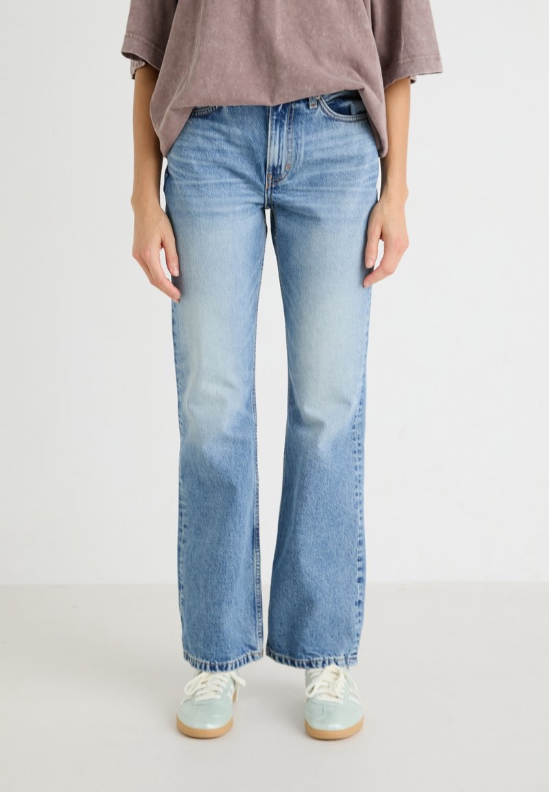 Personne portant un jean droit bleu clair, un haut marron ample et des baskets bleu clair avec des lacets blancs, debout devant un fond blanc uni.