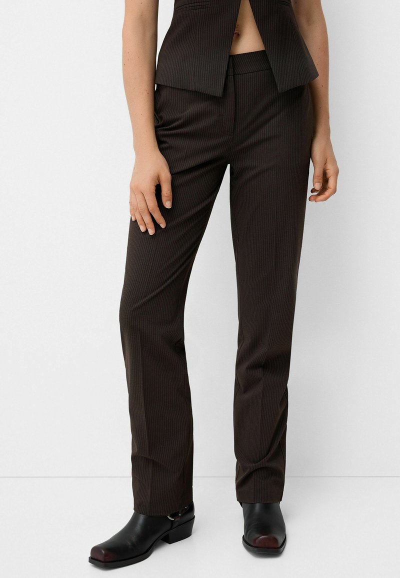 Pantaloni marroni a righe fitte con una vestibilità su misura, dotati di tasche laterali e una texture liscia. Indossati con stivali neri con tacco.