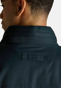 Detailný záber krku a hornej časti chrbta osoby v tmavom saku s prišitou nálepkou "JOULES" na oblasti goliera.