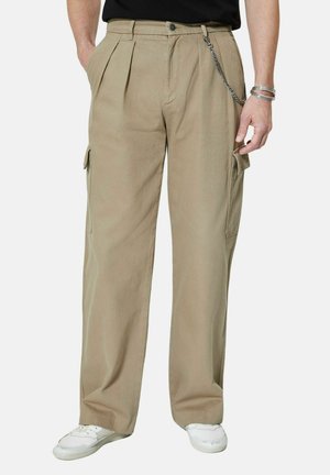 Pantalon cargo kaki à poches latérales, plis à la taille, accessoire chaîne, porté avec des baskets blanches et une chemise noire.