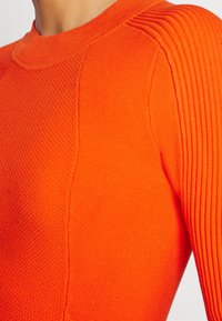 Gros plan d'un pull tricoté orange vif montrant un détail côtelé à l'épaule et un panneau avant texturé sur le haut du torse d'une personne.
