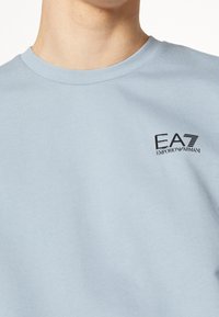 Ljust blå t-shirt med rund hals och svart EA7 Emporio Armani-logga på vänster bröst, bärs av en person som visas från nacken till midjan.