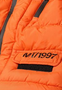 Siltam šūtu oranžu pūkainu jaku ar melnu Velcro plāksteri, uz kura uzrakstīts "MT/1997", un ar rāvējslēdzēja kabatu, kurai ir tumši pelēka aparatūra.
