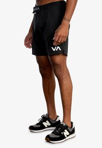 Svarta sportshorts med platt midjeband, logodetalj i vitt och sidoflikar för rörlighet. Bärs med svarta New Balance-sneakers.