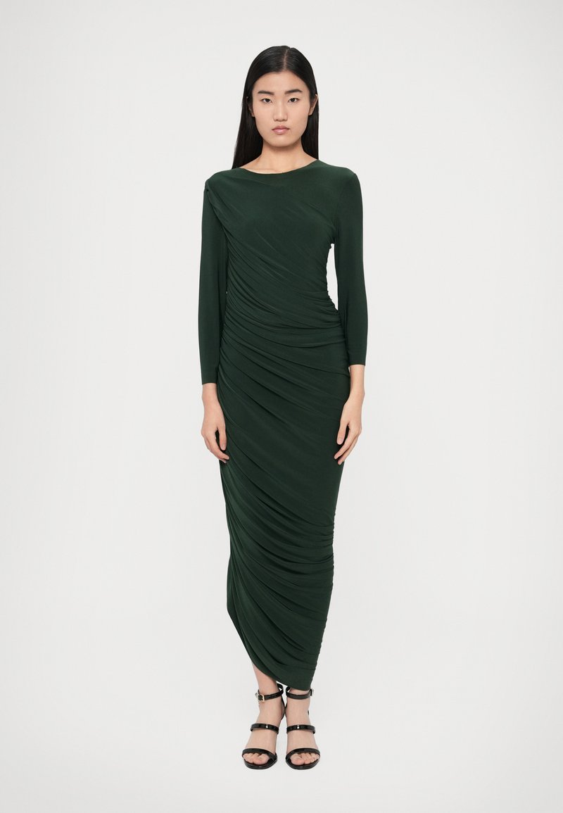 Sukienka z długim rękawem w ciemnozielonym kolorze z drapowanym designem, plisami i dekoltem w łódkę; w zestawieniu z czarnymi sandałami na paski.