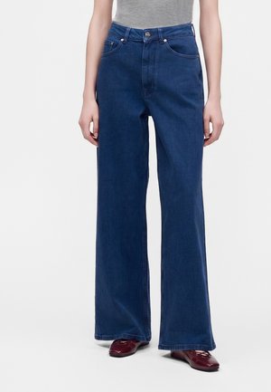 Kvinde iført mørkeblå jeans med høj talje og vidde ben med forlommer og bordeaux flade sko, stående foran en ensfarvet baggrund.
