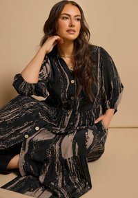 Schwarz-beige gemustertes Kleid mit durchgehender Knopfleiste, gepufften Ärmeln und gestuftem Rock. Weicher Stoff mit fließender Silhouette.