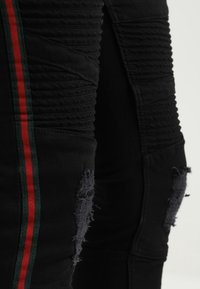 Detalle de unos pantalones vaqueros negros desgastados con costuras en relieve y una franja lateral verde-roja-verde.