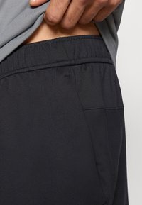 Fekete atlétikai rövidnadrág elasztikus derékpánttal, sima textúrával és finom varrási részletekkel. Részben látható szürke ing felette.