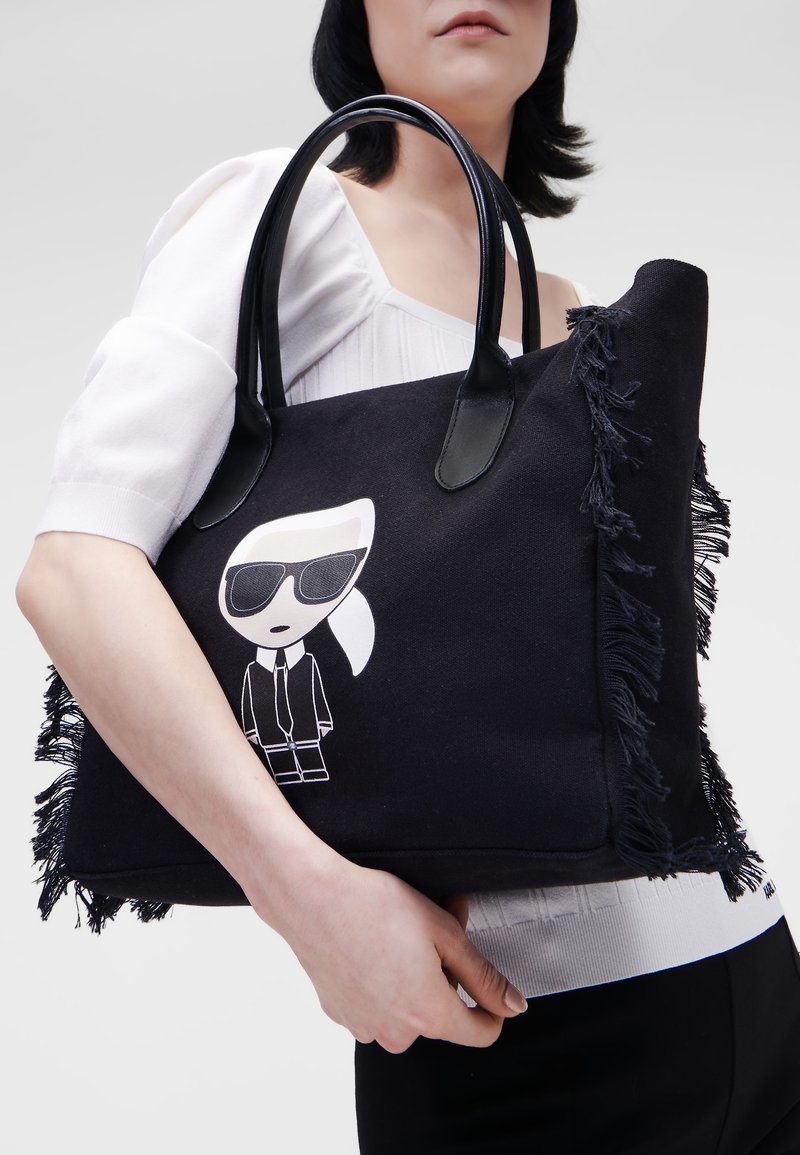 Femme portant un haut blanc tenant un grand sac cabas noir avec un personnage de dessin animé portant des lunettes de soleil et des franges sur les côtés.