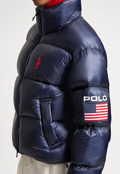 Navy puffer jacket with a high collar, red embroidered logo, side pockets, and a patch featuring "POLO" and an American flag on the sleeve.
