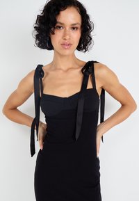 Μαύρο φόρεμα με εφαρμοστή σιλουέτα, που διαθέτει δομημένο μπούστο και μακριές σατέν κορδέλες στους ώμους. Απαλή, ελαστική ύφανση.