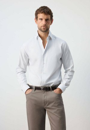 Mann mit kurzen braunen Haaren und Bart, der ein weißes Hemd mit Knöpfen und beige Hose trägt, steht mit den Händen in den Taschen vor einem einfarbigen Hintergrund.