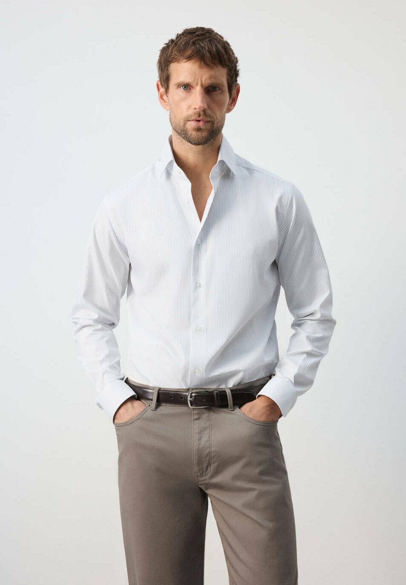 Homme aux cheveux courts et bruns avec une barbe, portant une chemise blanche boutonnée et un pantalon beige, debout les mains dans les poches devant un fond uni.