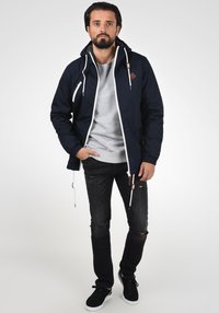 Marine vanntett jakke med hette, hvit glidelås og beige detaljer. Kombinert med en grå hettegenser, slitte svarte jeans og svarte joggesko.