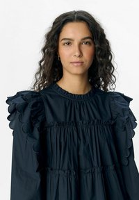 Schwarzes Kleid mit einem gerafften, gewellten Ausschnitt und Schultern, gestuften Rüschen und einer weichen Textur. Lange Ärmel vorhanden.