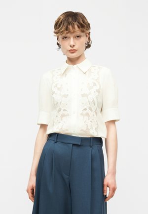 Žena s krátkými zvlněnými vlasy nosí bílou vyšívanou krátkosrstou halenku zastrčenou do tmavě modrých kalhot s vysokým pasem, stojí na bílém pozadí.