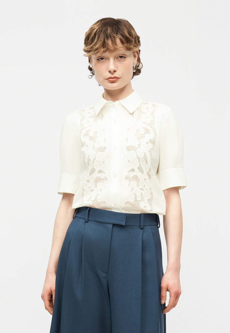 Γυναίκα με κοντά κυματιστά μαλλιά φοράει άσπρο κέντημα μπλουζάκι με κοντά μανίκια που είναι μέσα σε ψηλόμεσο σκούρο μπλε παντελόνι, στέκεται μπροστά σε λευκό φόντο.