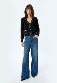 Svart stickad cardigan med v-ringning, knappfram och vita sömnadsdetaljer. Kombinerad med ljusblå vida jeans. Slät textur.