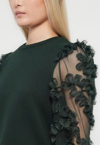 Donna che indossa un top verde scuro con maniche floreali trasparenti e arricciate, mostrata dalle spalle al mento.