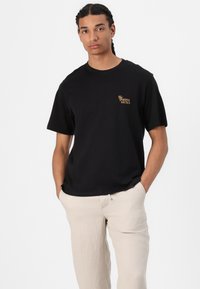 T-shirt en coton noir à manches courtes, avec un petit logo en texte doré sur la poitrine gauche. Le modèle porte un pantalon beige clair.