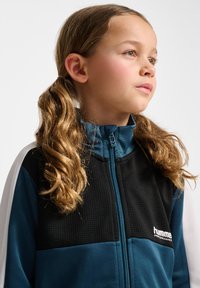 Teal en zwarte ritsjack met textuurstof, witte accenten en een gelabeld logo. Sluiting bij de kraag; glad, sportief ontwerp.