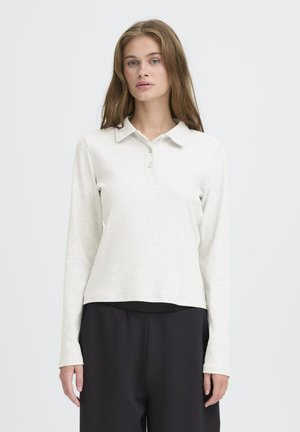 Junge Frau mit langen Haaren, die ein hellbeiges Langarm-Polo-Shirt und schwarze Hosen trägt und vor einem schlichten weißen Hintergrund steht.