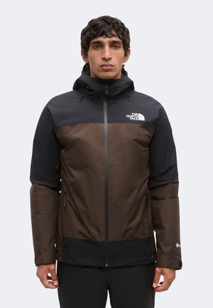 Άνδρας που φοράει καφέ και μαύρο μπουφάν North Face με κουκούλα, στέκεται με το πρόσωπο προς τα εμπρός σε απλό ανοιχτό φόντο.