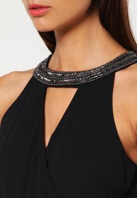Kvinna som bär en svart ärmlös klänning med en pärl- och paljettdetalj vid kragen och en V-formad utskärning.